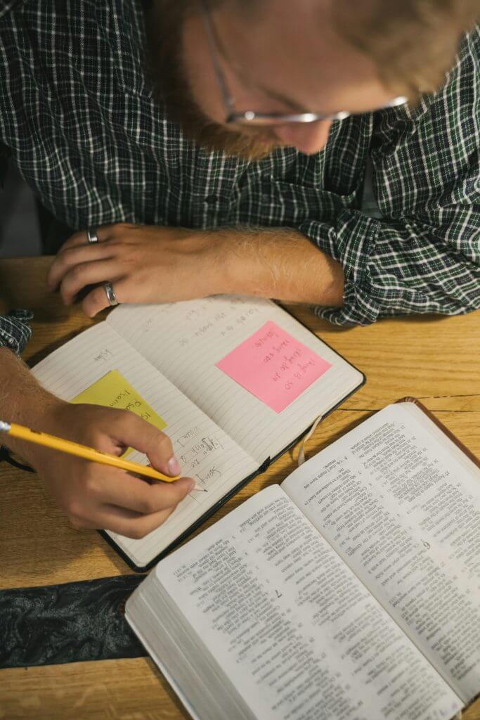 A man writes notes in a notebook while reading an open Bible on a wooden table.