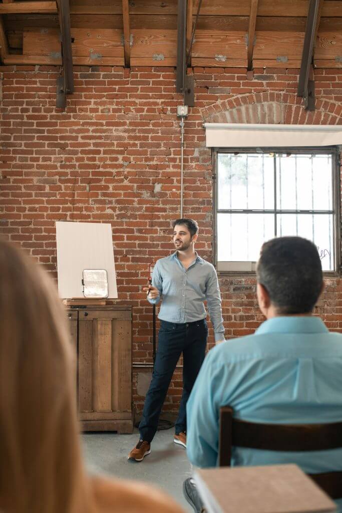 A young man stands in front of a class presenting, with a brick wall backdrop and attentive audience.