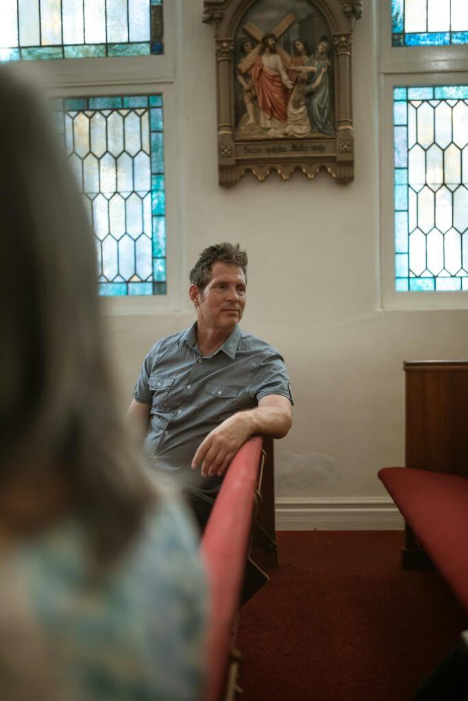 A contemplative man sits in a church pew with stained glass windows and religious art.